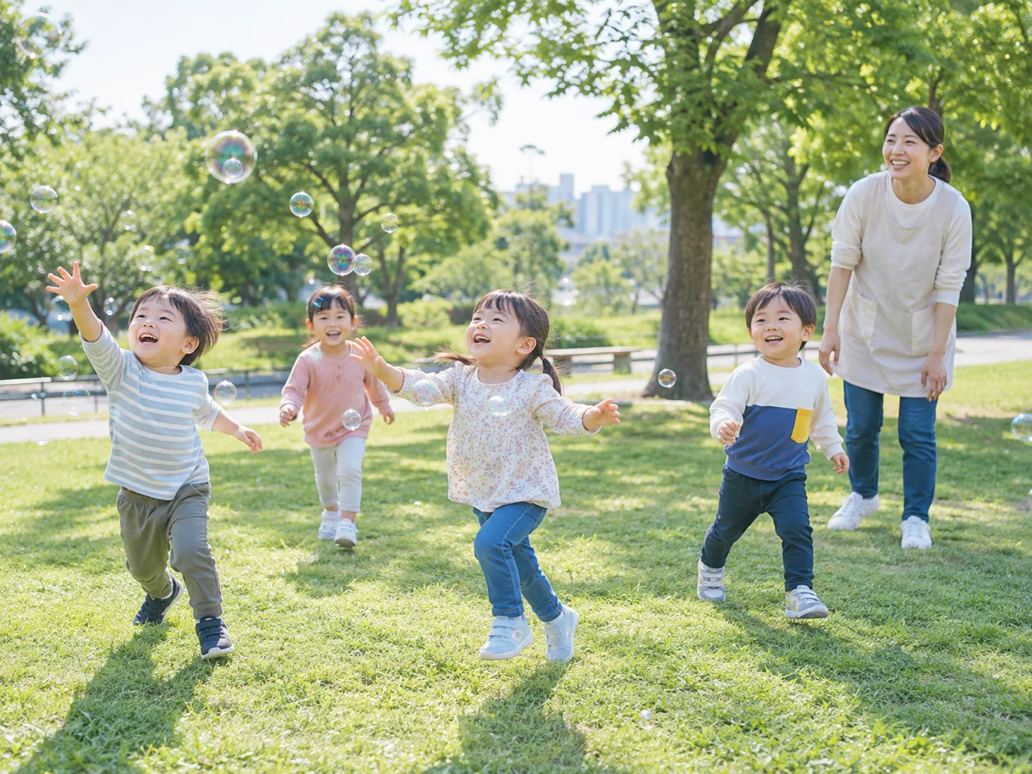 芝生の公園でシャボン玉を追う子どもたちと見守る先生のイメージ
